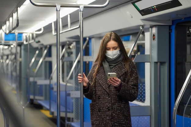 Uma mulher com uma máscara médica para evitar a propagação do coronavírus está de pé e usando um smartphone em um vagão vazio do metrô