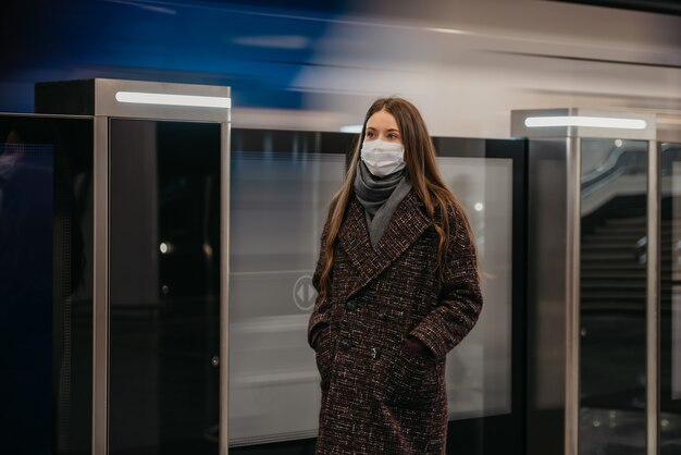 Uma mulher com uma máscara médica para evitar a propagação do coronavírus está de pé perto do trem que parte na plataforma do metrô. uma garota com máscara cirúrgica está mantendo distância social em uma estação de metrô.