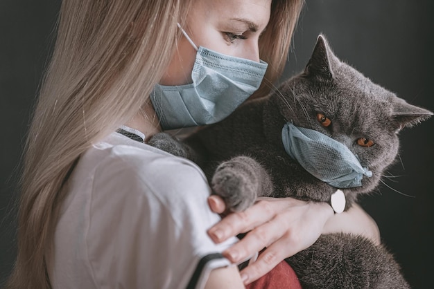 Foto uma mulher com uma máscara médica segura um gato britânico nos braços, o gato também está com uma máscara médica