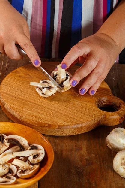 Uma mulher corta cogumelos champignon com uma faca em uma placa de madeira