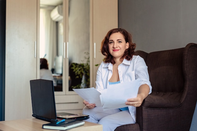 Uma mulher de 30 anos, com cabelos escuros e camisa branca, senta-se em uma poltrona em uma pequena mesa com um laptop, segura o papel nas mãos e trabalha remotamente em seu escritório em casa.