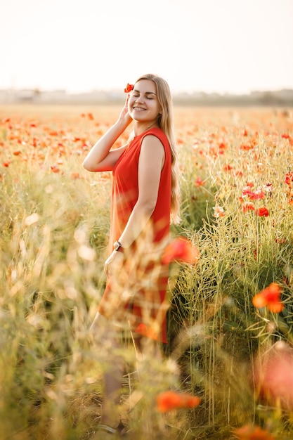 Uma mulher de aparência europeia com longos cabelos loiros e um vestido vermelho de verão, ela está em um campo de papoulas florescendo