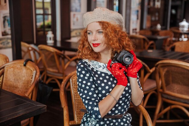 Uma mulher de boina e vestido vintage segura uma câmera nas mãos e tira fotos