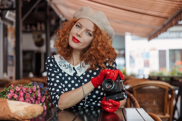 Uma mulher de boina e vestido vintage segura uma câmera nas mãos e tira fotos