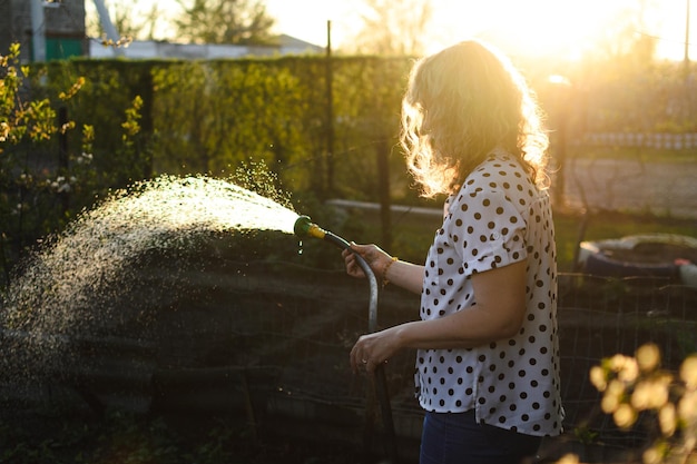 Uma mulher de cabelo encaracolado à noite rega o jardim com água de uma mangueira com um pulverizador