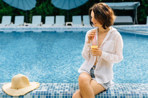 Uma mulher de camisa bebe suco espremido na hora de um copo enquanto está sentada na beira da piscina durante suas férias de verão ou relaxando à beira da água Garota latina cura com suco