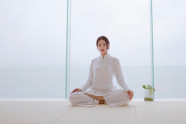 Uma mulher de camisa branca está sentada em uma pose de ioga em frente a uma janela com vista para o mar.