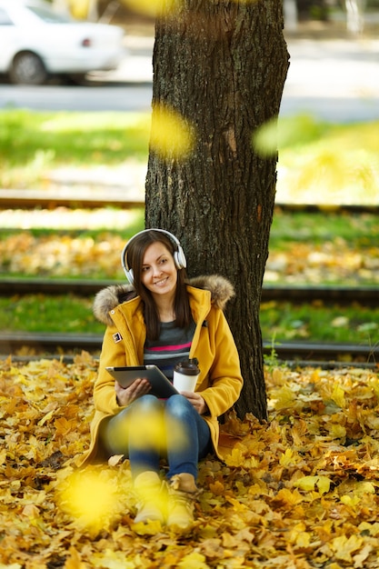 Uma mulher de casaco amarelo e calça jeans sentada com uma xícara de café ou chá e ouvindo música debaixo de uma árvore com um tablet nas mãos e fones de ouvido no outono parque da cidade em um dia quente. Folhas de outono dourado.