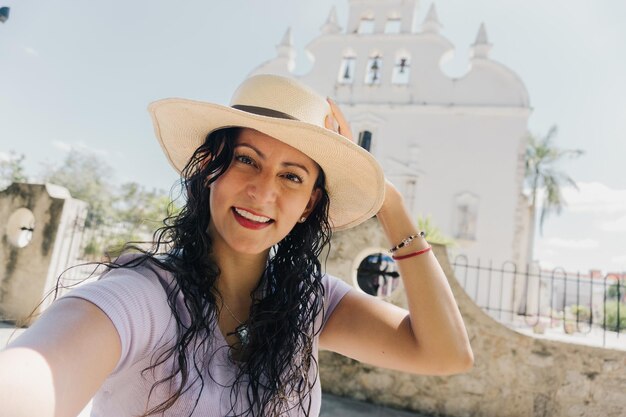 Uma mulher de chapéu branco tira uma selfie em frente a um prédio branco.