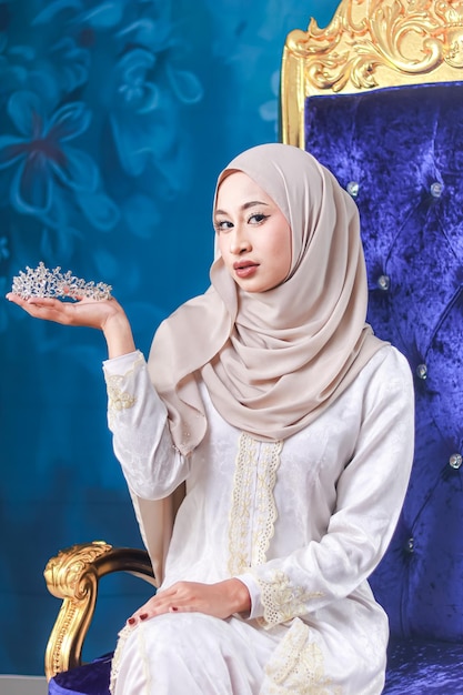 Uma mulher de hijab segura uma coroa em frente a uma parede azul.