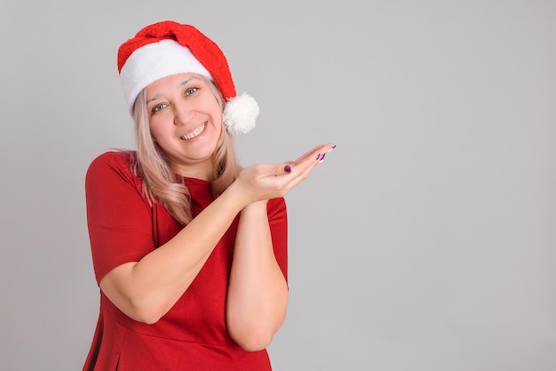 Uma mulher de meia-idade de 40 anos com um chapéu de Papai Noel aponta o dedo para copyspace sobre um fundo cinza.