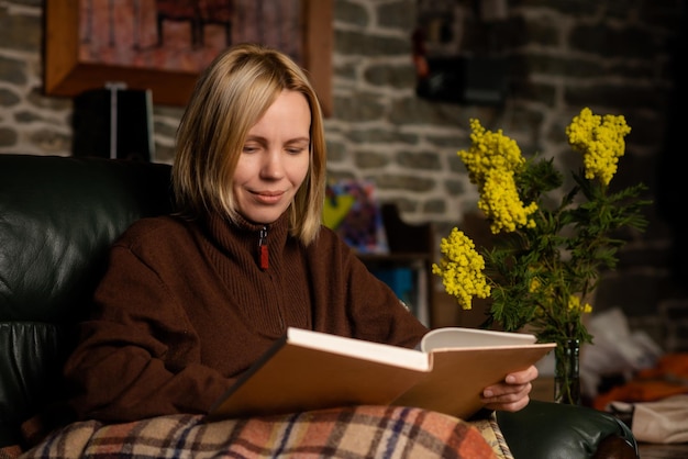 Uma mulher de meia-idade de quarenta anos está lendo um livro no sofá Mulher com roupas quentes coberta com um cobertor Fotografia escura