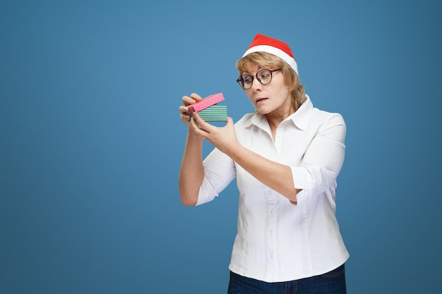 Uma mulher de meia-idade em uma camisa branca, usando um chapéu de Natal em um fundo azul. Uma mulher segura uma caixa de presente.