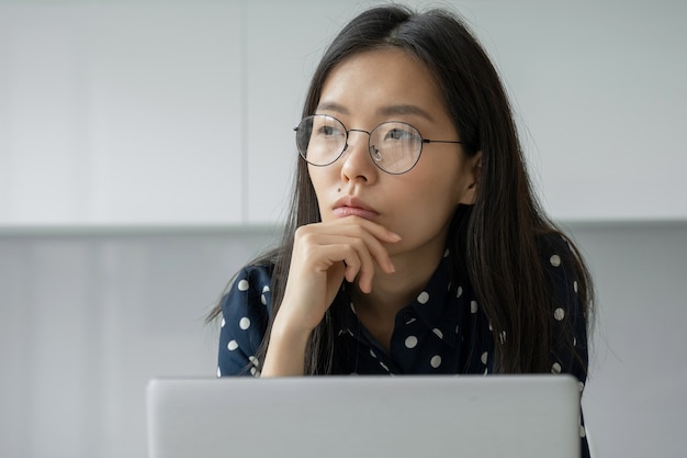 Uma mulher de negócios asiática pensativa com óculos e trabalhando com um laptop olha para a distância