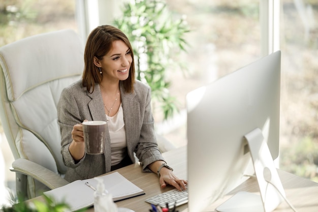 Uma mulher de negócios atraente bebendo café enquanto estiver usando um computador em seu escritório em casa.