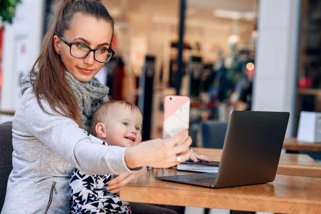 Uma mulher de negócios bem sucedido trabalha em um laptop com sua filha no local de trabalho. uma mulher bonita faz uma selphie com seu bebê sorridente.
