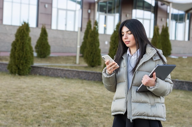 Uma mulher de negócios em uma jaqueta elegante olha para informações em um telefone celular e segura um laptop em suas mãos no contexto de edifícios comerciais