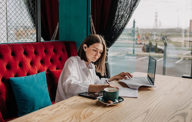 Uma mulher de negócios focada em fones de ouvido sem fio está sentada à mesa em um café e trabalhando com um laptop