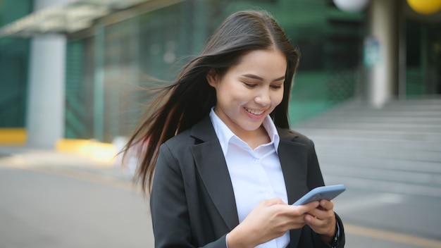 Uma mulher de negócios jovem vestindo terno preto está usando o telefone inteligente, na cidade, o conceito de estilo de vida de negócios.