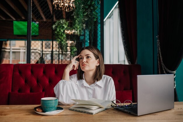 Uma mulher de negócios morena atenciosa com uma blusa branca está sentada à mesa em um café com um telefone e um laptop