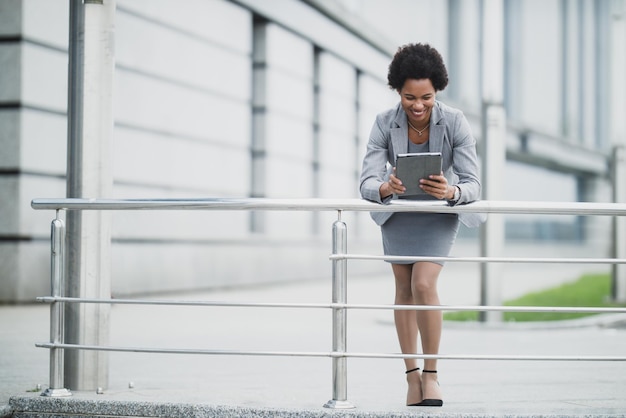 Uma mulher de negócios negra alegre trabalhando em um tablet digital em frente a um prédio corporativo.