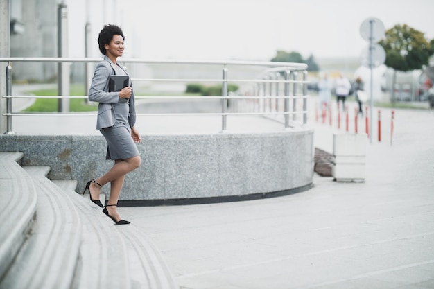 Uma mulher de negócios negra sorridente enquanto descia as escadas em frente a um prédio corporativo.