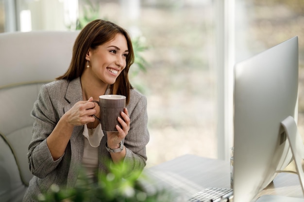 Uma mulher de negócios sorridente, desfrutando de uma xícara de café enquanto trabalhava no computador no escritório em casa.