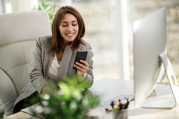 Uma mulher de negócios sorridente sentada sozinha em seu escritório em casa e usando smartphone.