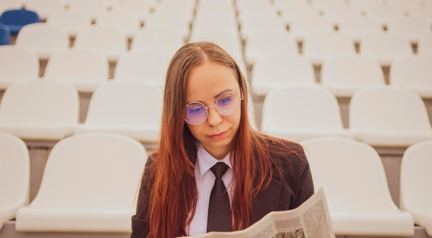 Uma mulher de óculos e um terno de negócios lê um jornal enquanto está sentado no estádio