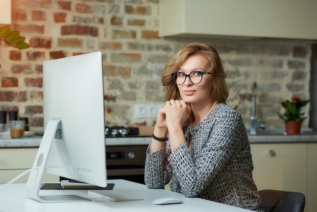 Uma mulher de óculos trabalha remotamente em um computador desktop em seu estúdio. Uma chefe mulher senta os braços cruzados e pensando durante uma videoconferência em casa.