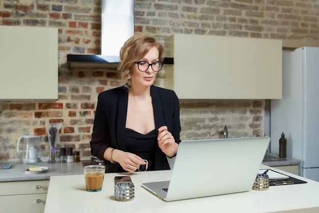 Uma mulher de óculos trabalha remotamente em um laptop na cozinha dela. Uma garota discute com seus colegas em um briefing de negócios on-line em casa. .