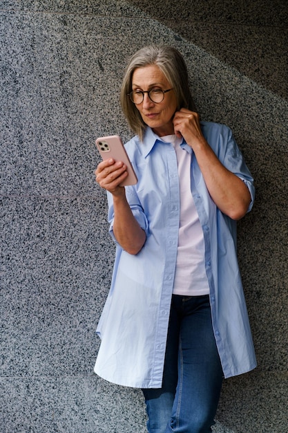 Foto uma mulher de pé contra uma parede enquanto segura um telefone celular na mão olhando para a tela