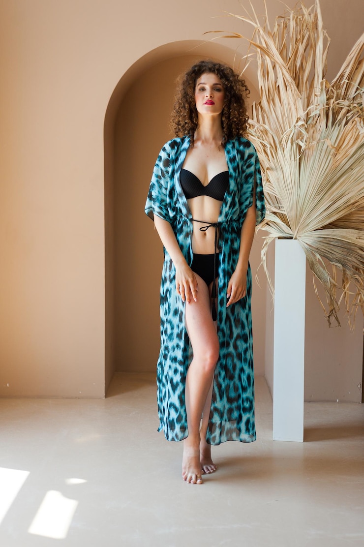 Foto uma mulher de quimono azul fica em frente a uma parede com uma planta ao fundo.