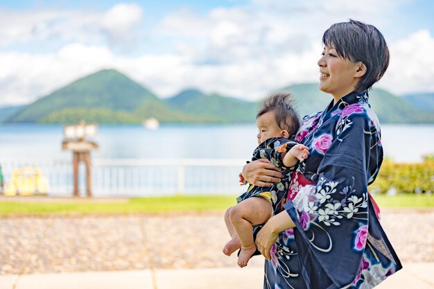 Uma mulher de quimono segura um bebê em frente a um lago.