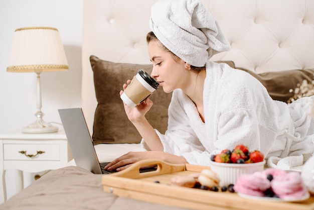 Uma mulher de roupão de banho e com uma toalha na cabeça depois de um banho na cama com um laptop trabalhando e aprendendo. Trabalho online em casa. Fique em casa durante a quarentena