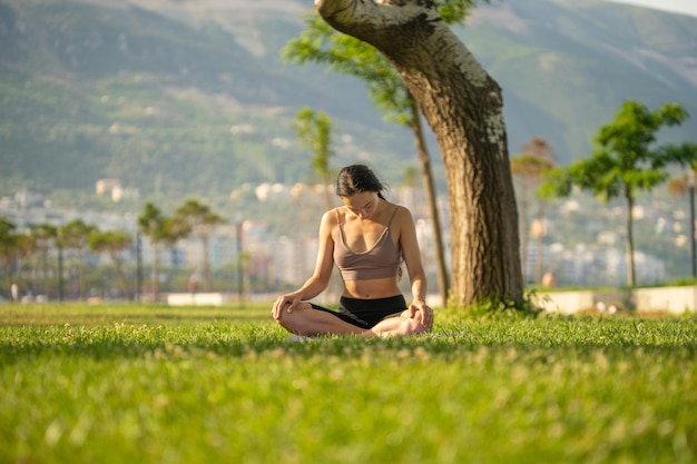 Uma mulher de sutiã esportivo marrom e short preto sentado no tapete de ioga fazendo meditação com a mão junto um