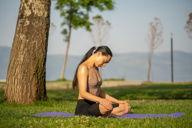 Uma mulher de sutiã esportivo marrom e short preto sentado no tapete de ioga fazendo meditação com a mão junto um