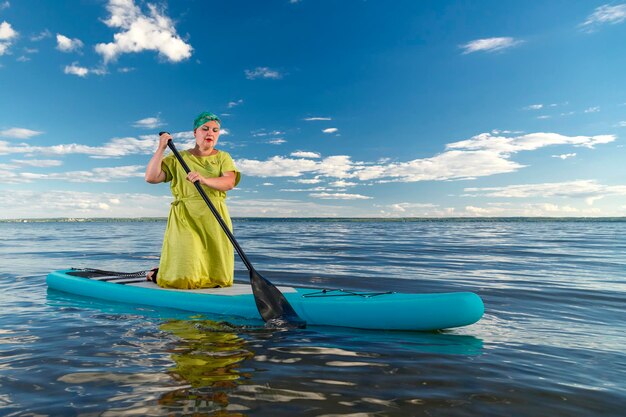 Uma mulher de vestido e um cocar de pé em uma prancha de SUP com um remo flutua na água contra o céu azul