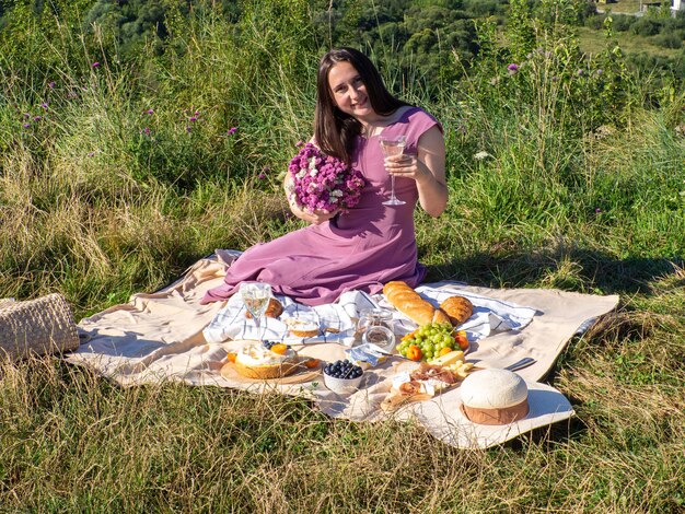 Uma mulher de vestido rosa e com cabelos escuros sentada em um cobertor bege segurando um copo e bebendo vinho branco. Conceito de fazer piquenique em um parque da cidade durante as férias de verão ou fins de semana. Flores cor-de-rosa.