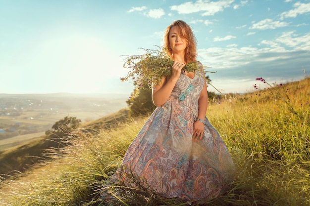 Uma mulher de vestido segura um ramo de flores em frente a uma colina.