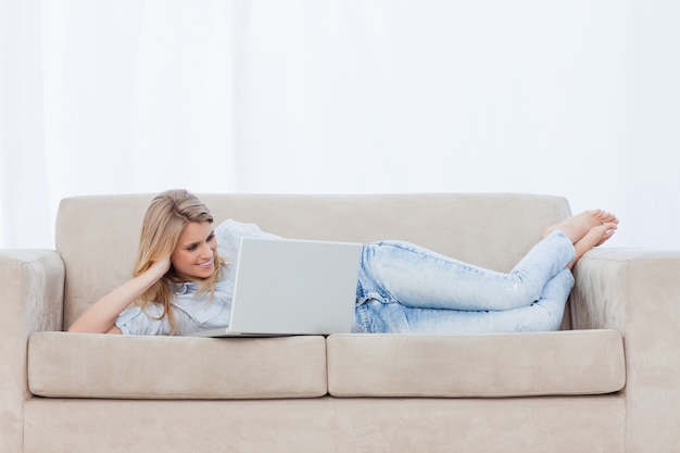 Uma mulher deitada em um sofá descansando a cabeça na mão está usando seu laptop