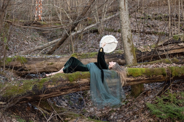 Uma mulher deitada em um tronco de árvore toca um tambor xamânico em um dia de primavera