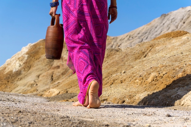 Uma mulher descalça com roupas étnicas carrega na mão um jarro de barro