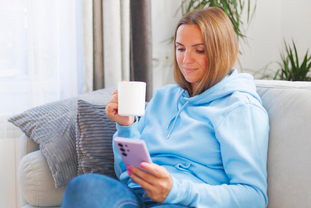 Uma mulher descansa em um sofá confortável smartphone na mão e uma chávena de chá