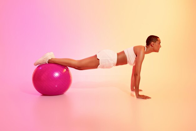 Uma mulher disciplinada com a cabeça rapada ocupa uma posição de prancha em uma bola de exercício rosa