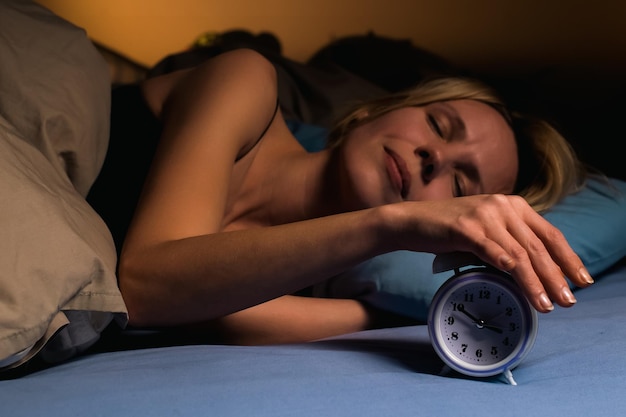 Uma mulher dorme na cama há um despertador por perto Sono saudável descanso conceito de ritmo circadiano Uma mulher de quarenta anos de idade em uma cama azul escura