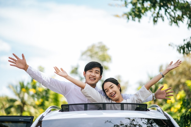 Uma mulher e um homem estão em um carro com o teto solar aberto e os braços estendidos.