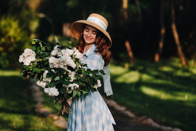 Uma mulher elegante com um chapéu de palha posa em flores lilás em um parque ensolarado de primavera. Retrato calmo de uma linda garota em pé com um buquê de lilases no jardim primavera.