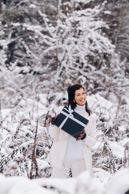 Uma mulher elegante com um terno branco com um presente de ano novo nas mãos em uma floresta de inverno Uma garota na natureza em uma floresta de neve com uma caixa de presente