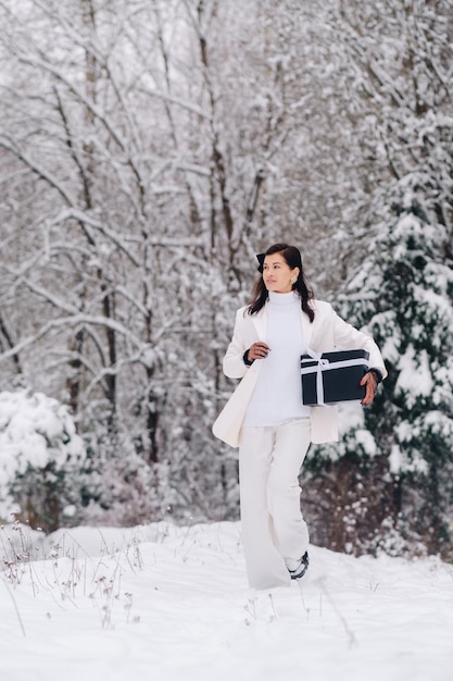 Uma mulher elegante com um terno branco com um presente de ano novo nas mãos em uma floresta de inverno Uma garota na natureza em uma floresta de neve com uma caixa de presente